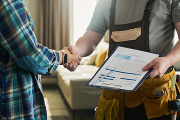 Caucasian middle aged man wearing tool belt holding invoice clipboard shaking hands with unseen...