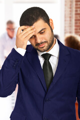 Male professional in business attire touching his forehead in a moment of stress or worry. Office background highlights a workplace setting, portraying themes of career challenges and professional str