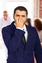 A man in a business suit looking nervous and biting his nails, with office coworkers in the background, depicting anxiety or stress in a professional environment.