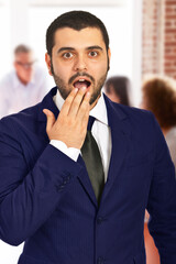 Surprised businessman covering mouth with his hand in an office environment. A bright and professional setup showcasing a shocked reaction, capturing a candid and engaging moment.