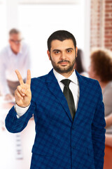 Professional individual in business attire gesturing peace symbol, evoking positivity and confidence, set against a blurred office background representing teamwork and professional engagement.
