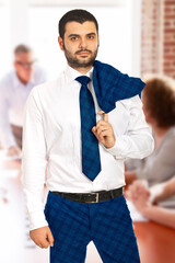 A professional man dressed in business attire stands confidently in a modern office environment. Background shows colleagues collaborating, symbolizing teamwork and corporate atmosphere.