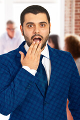 Shocked adult male in a blue suit, indoors with blurred people in the background. Expressing a surprised expression while wearing formal attire, reflecting emotional reaction and professional atmosphe