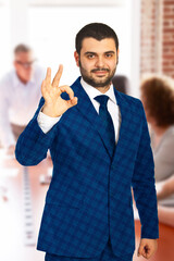 Confident male professional in a suit giving an OK hand gesture in a workplace. The image conveys confidence, approval, and a positive attitude, ideal for business presentations or motivational concep