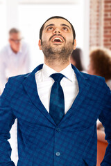 A professional man in a blue suit expresses joy and satisfaction in a dynamic office environment, highlighting success and accomplishment. The background shows a collaborative team working together, e