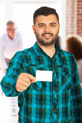 A man in a plaid shirt holding a blank card, while people converse in the background.