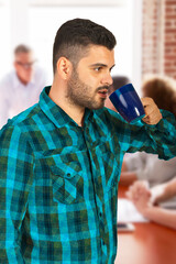 A young man wearing a plaid shirt enjoys a hot beverage during a casual team meeting. Background shows indistinct coworkers collaborating, reflecting relaxation and focus. Perfect for themes of teamwo