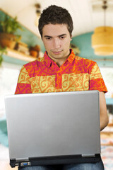 A focused young man wearing a colorful shirt works on his laptop in a lively room, surrounded by plants and a bright atmosphere, indicating productivity and a positive indoor environment.