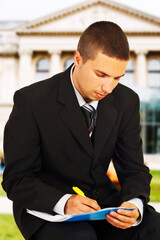 A young business professional is diligently writing notes while seated outside a stately building.