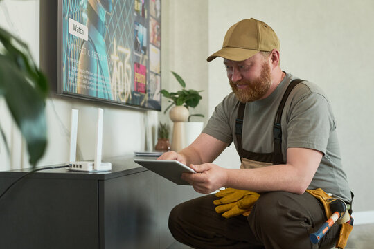 Caucasian middle aged man with beard wearing cap setting up wireless router using digital tablet in modern living room, focusing on configuring home internet connection