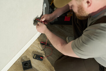 Caucasian middle aged man kneeling on wooden floor testing electrical outlet with digital multimeter, holding probes near socket, performing maintenance or repair work
