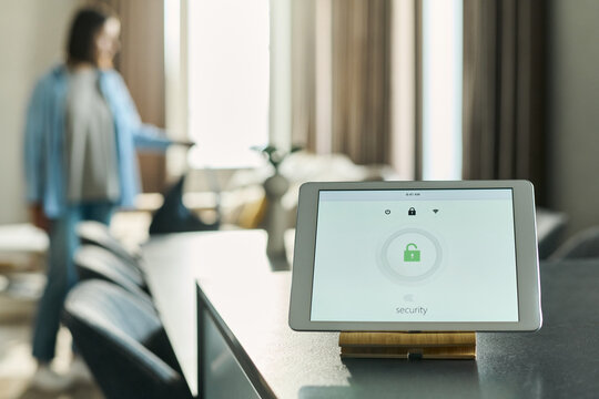 Tablet displaying security system interface with lock icon on kitchen counter, Caucasian young adult woman standing in background adjusting object, home interior visible