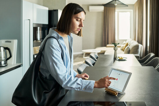 Young adult Caucasian woman standing in modern kitchen interacting with smart home control panel on countertop, carrying large shoulder bag, focusing on touchscreen interface - Powered by Adobe