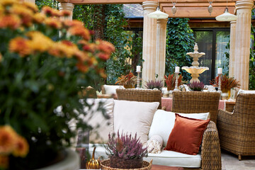 Cozy outdoor patio with wicker furniture, decorative columns, fountain and autumn floral arrangements. Elegant rustic style terrace with natural light and warm seasonal atmosphere