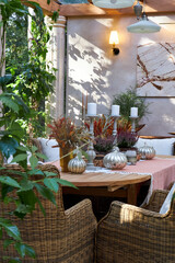 Elegant autumn table setting on a terrace with wicker chairs, candles, silver pumpkins and seasonal floral arrangements. Cozy rustic atmosphere with natural light and greenery