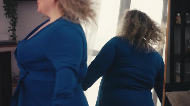 Beautiful curvy woman in blue dress posing near mirror, showing body positivity
