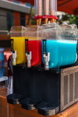 Three beverage dispensers showcase vibrant yellow, red, and blue drinks at a lively outdoor gathering. The festive atmosphere is enhanced by the colorful drinks, ready to quench thirst. Ice slush.
