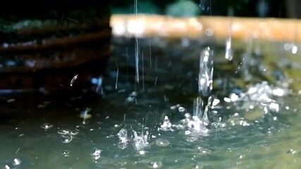 Close-up of water droplets splashing into a rustic stone fountain bowl in a garden, creating ripples and reflections with greenery in the background - Powered by Adobe