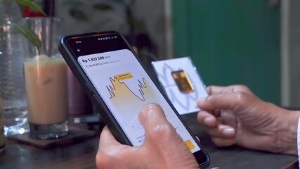 A footage of Person using a smartphone to check investment balance while holding a gold bar, representing online finance, savings, and digital economy - Powered by Adobe