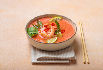 Creamy flavourful tom yum soup, a popular dish of Thailand, is served in a bowl, garnished with fresh shrimp, lime wedges, herbs, and chili peppers, accompanied by chopsticks on a linen napkin