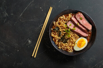 Bowl of ramen noodle soup with sliced beef, a boiled egg, seaweed, and green onions, served with chopsticks on a dark background, asian style soup top view, copy space