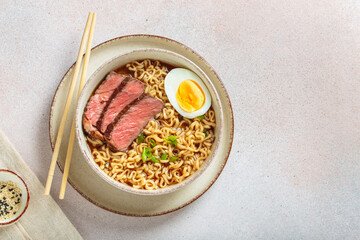 Bowl filled with ramen noodles, tender sliced beef, boiled egg and fresh green onions, asian style soup on light beige background top view, copy space