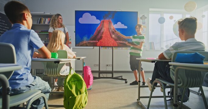 Elementary School Boy with Tablet Computer Presenting Homework, Explaining Volcanic Eruption Process in Front of Classmates and Teacher Using Digital Screen. Smart Kids Studying Geology in Classroom.