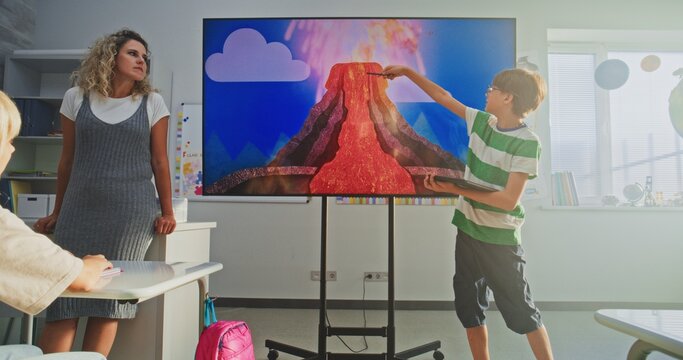 Elementary School Boy with Tablet Computer Presenting Homework, Explaining Volcanic Eruption Process in Front of Classmates and Teacher Using Digital Screen. Smart Kids Studying Geology in Classroom.