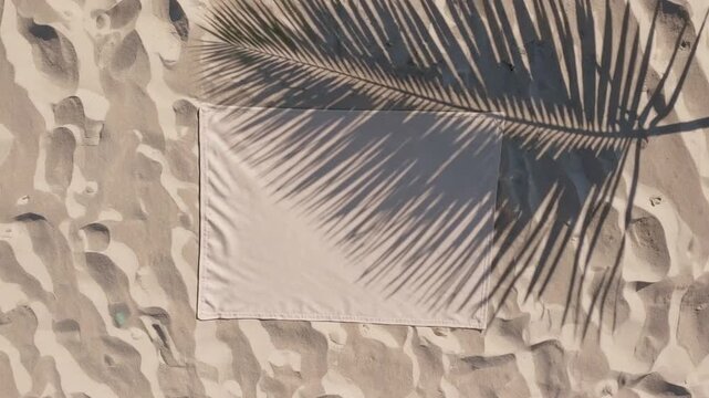 summertime backdrop captured from aerial perspective, showcasing beach towel laid out upon the sandy shore, with the distinctive shadow pattern of palm frond cast by the sun, embodying minimalist aes