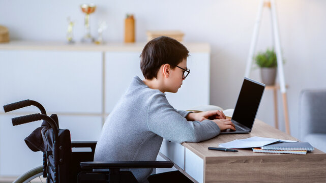 Side view of focused teen boy in wheelchair using laptop for online studies or communication with teacher at home. Industrious disabled adolescent learning remotely, talking to his tutor