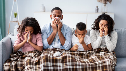 Flu And Illness Concept. Portrait of sick young African American family blowing noses with napkins together while sitting on the couch covered with blanket. Four unwell people with cold at home