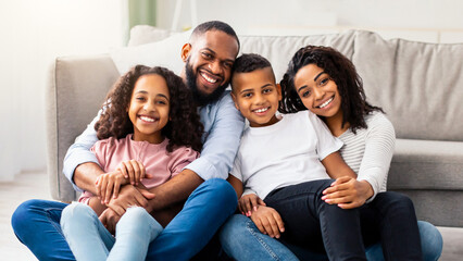 Happy Loving Family. Portrait of cheerful African American man and woman sitting on the floor carpet in living room, posing for photo, embracing smiling boy and girl. Four positive black people