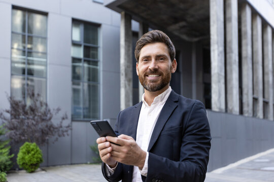 Smiling professional businessman holding a smartphone outside a modern office building