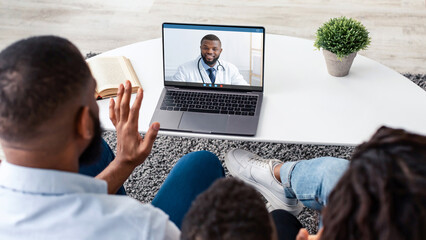 Over the shoulder view of young black family using pc laptop computer with blank empty screen for mock up template, sitting on the sofa in living room at home, waving to webcam, having videocall