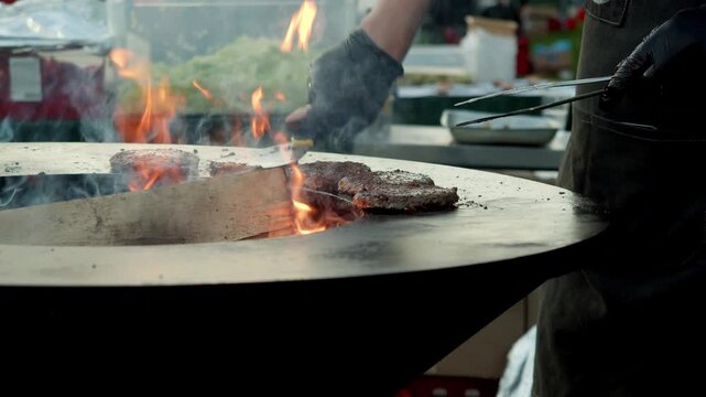 The chef employs metal tongs to turn the burgers on an open fire on a circular pit grill during the evening barbecue.