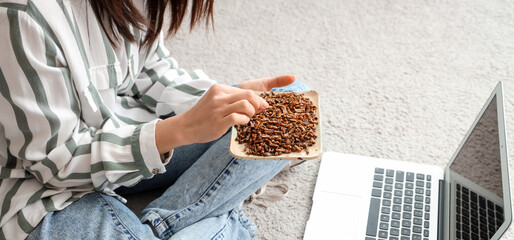 Young woman with fried insects and laptop sitting on carpet at home