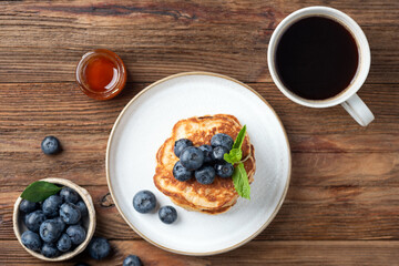 Pancakes with blueberries, honey and cup of black coffee on rustic wooden table background, top view