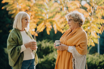 Fototapeta premium Two senior women talking and drinking coffee in autumn park