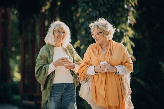 Two senior women smiling and walking in park, enjoying coffee and conversation - Powered by Adobe