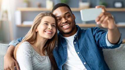 Happy Black Man And White Woman Taking Selfie On Smartphone Together At Home, Romantic Interracial Couple Embracing And Posing At Camera While Resting On Couch In Living Room, Closeup Shot