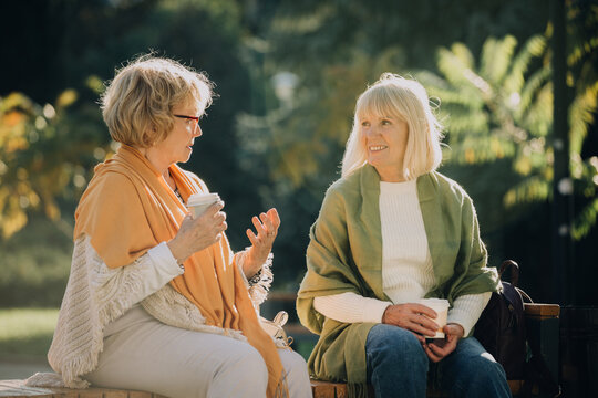 Two senior women talking and drinking coffee on a bench in a park