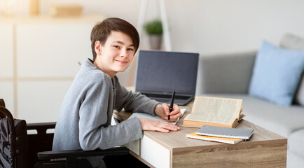 Online education for dsiabled teens. Happy teenage boy in wheelchair studying online from home, using laptop and textbooks, doing homework, having remote lesson, copy space