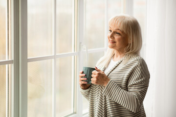 Calm elderly woman standing near and looking out of window in living room, drinking tea, thinking or dreaming, smiling mature grandmother enjoying coffee relaxing at home, free copy space