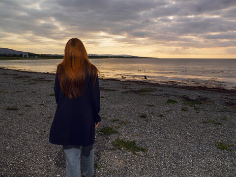 Red hair girl in black long coat is walking on a stone beach towards ocean at sunset wit dark and moody sky. Enjoy nature concept. Model in stylish glamour outfit.