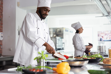 Black head chef chopping a fresh bell pepper in a modern workspace, preparing raw materials for a delicious gourmet recipe. Slicing veggies at the kitchen counter with utensils and cookware.