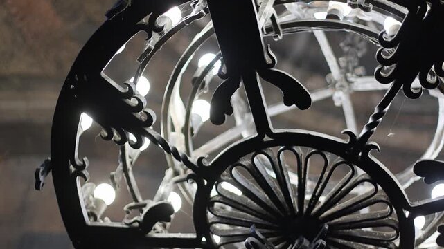 A detailed, close-up view of an antique black wrought iron chandelier with multiple bulbs, showcasing its intricate scrollwork and decorative design against a dimly lit, textured background.