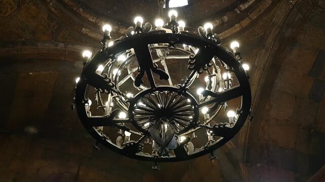 A detailed, close-up view of an antique black wrought iron chandelier with multiple bulbs, showcasing its intricate scrollwork and decorative design against a dimly lit, textured background.