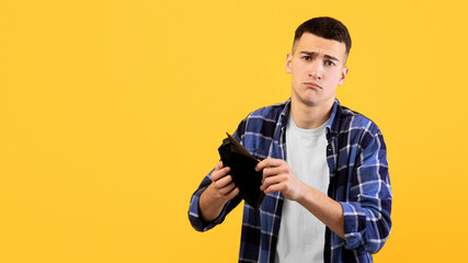Bankruptcy concept. Upset young man showing empty wallet, suffering from loss of money on orange studio background. Sad millennial guy losing finances, being unemployed during economic crisis