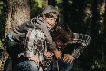 A boy in a hoodie hugs an older boy in a plaid shirt from behind in a forest, sharing a tender and playful moment in the sunlight.
