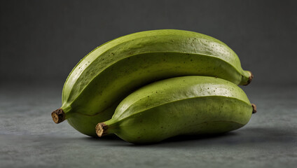 A close-up of a bunch of thick, green, unripe bananas or plantains. The fruit is positioned to show its size and full shape, with the light highlighting the deep color and smooth surface. The backgrou